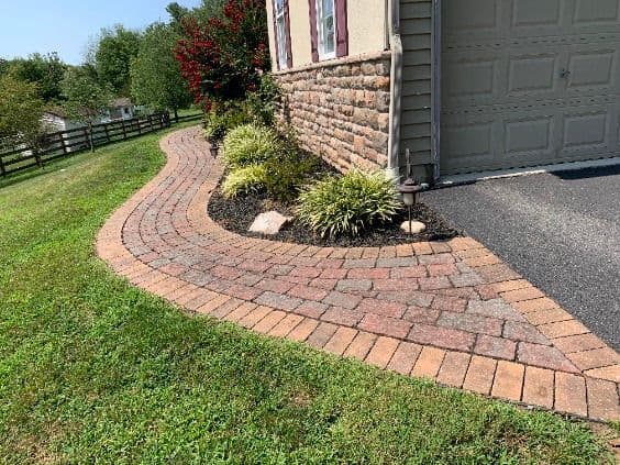 Curved brick walkway with landscaping near a house and garage, surrounded by greenery.